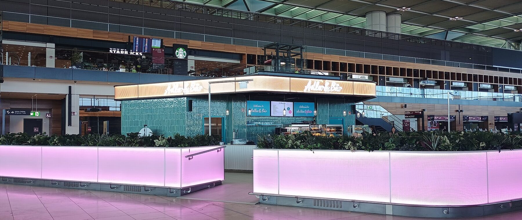 The check-in hall of BER Terminal 1, with a café counter lit in pink, a Starbucks on the gallery above and a red hanging artwork overhead