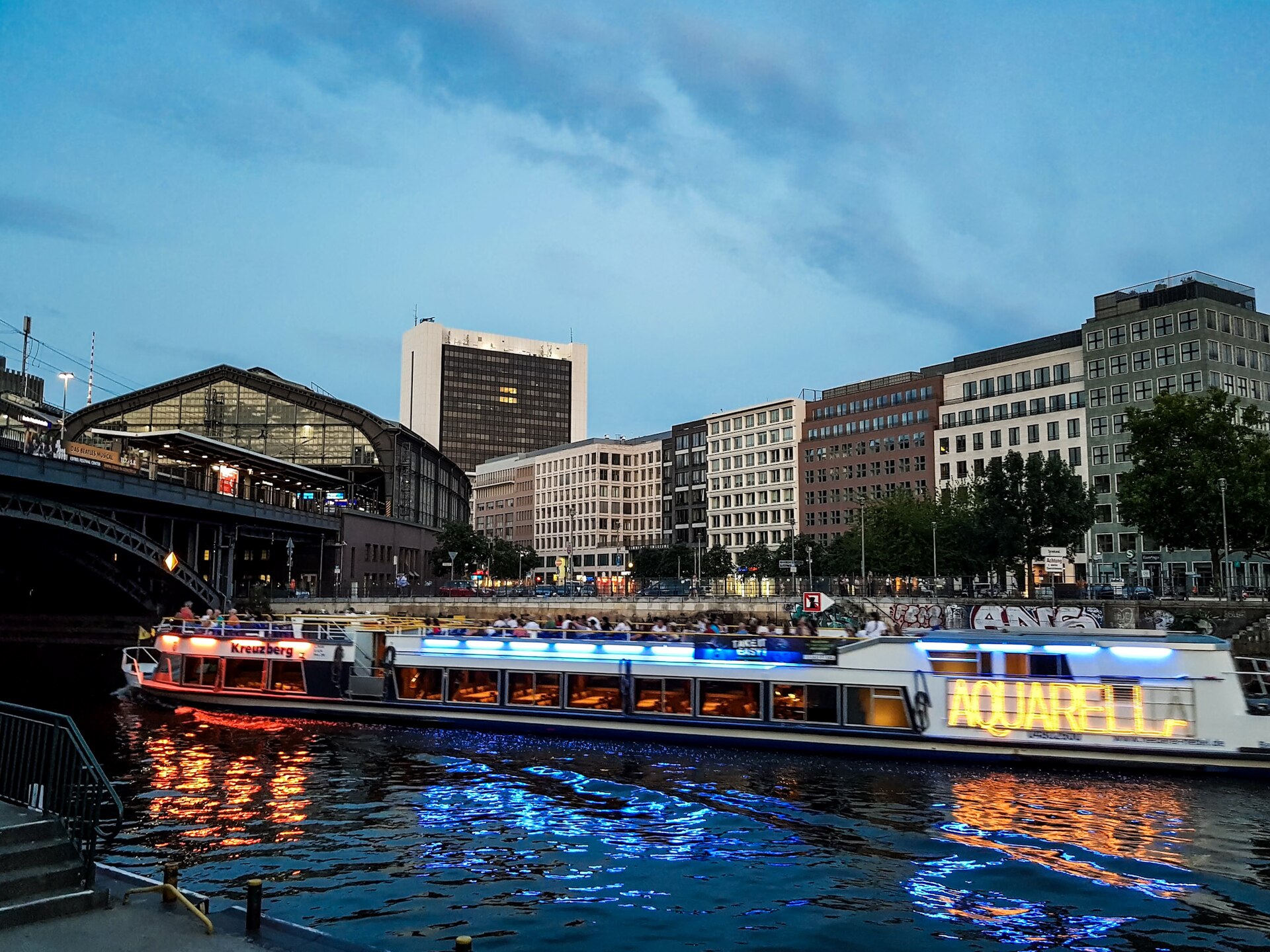 A tour boat lit with blue and orange lights on the Spree at dusk beside Friedrichstraße station, with office buildings behind