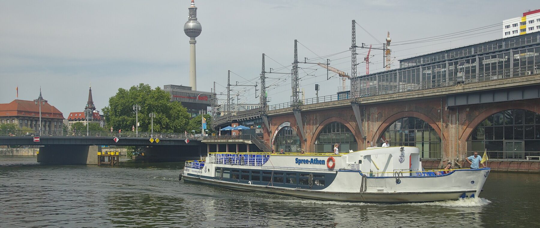 A white sightseeing boat on the Spree beside the arches of the Jannowitzbrücke railway bridge, with the TV Tower rising behind