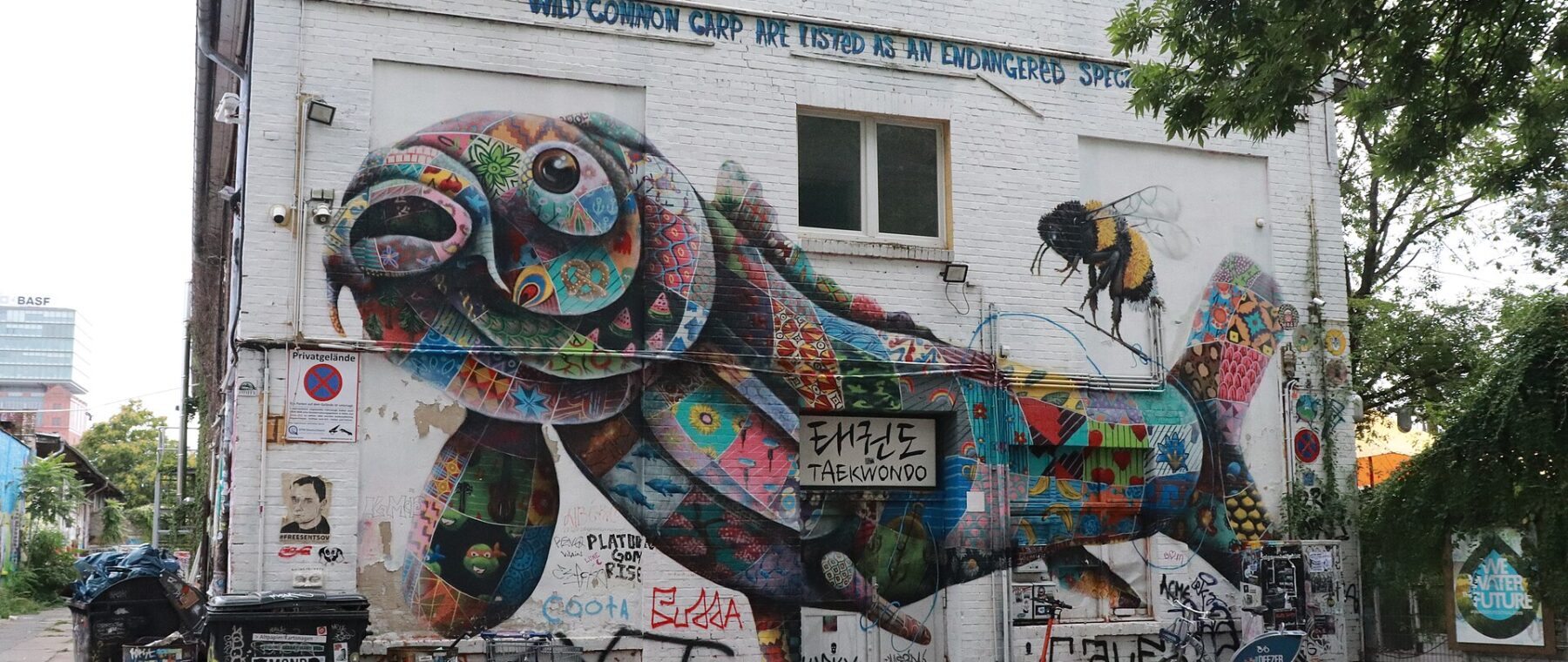 A huge multicoloured mural of a carp covering the side of a white building on the RAW-Gelände in Friedrichshain, with a painted bee beside it