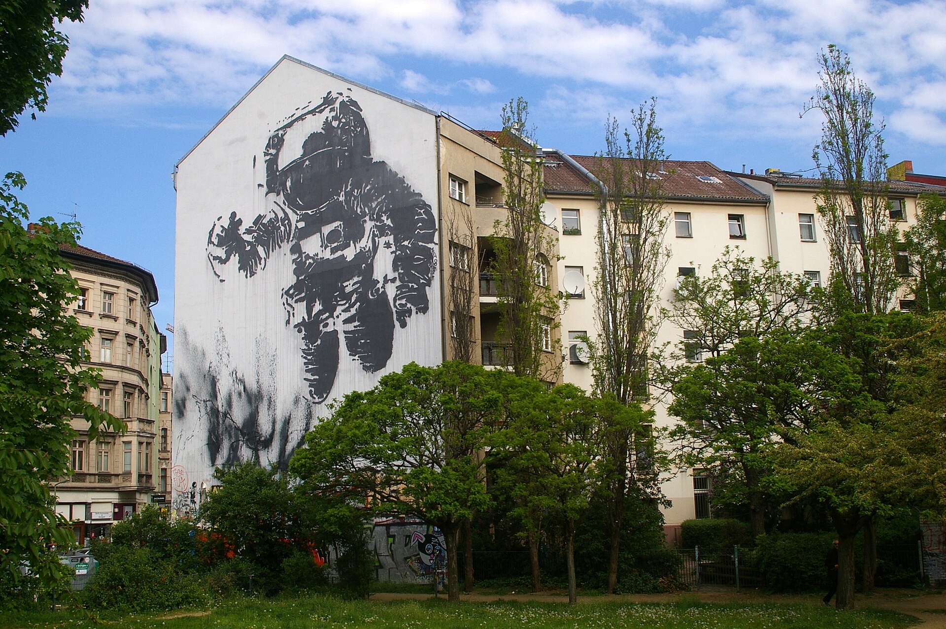 A huge black-and-white mural of a floating cosmonaut on the white gable wall of an apartment building in Kreuzberg, above trees and a small park