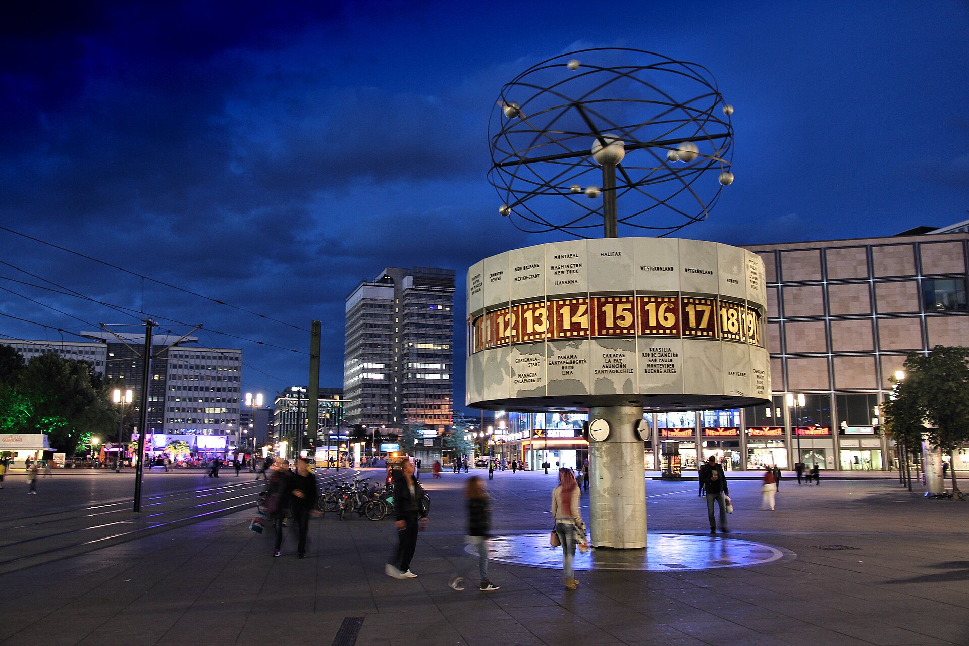 The Weltzeituhr world time clock on Alexanderplatz in Berlin lit up at dusk with people crossing the square