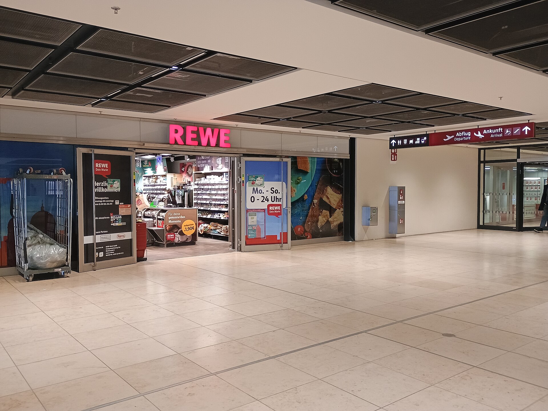 The entrance to a REWE supermarket inside BER Terminal 1, with a sign showing opening hours of Monday to Sunday 0 to 24